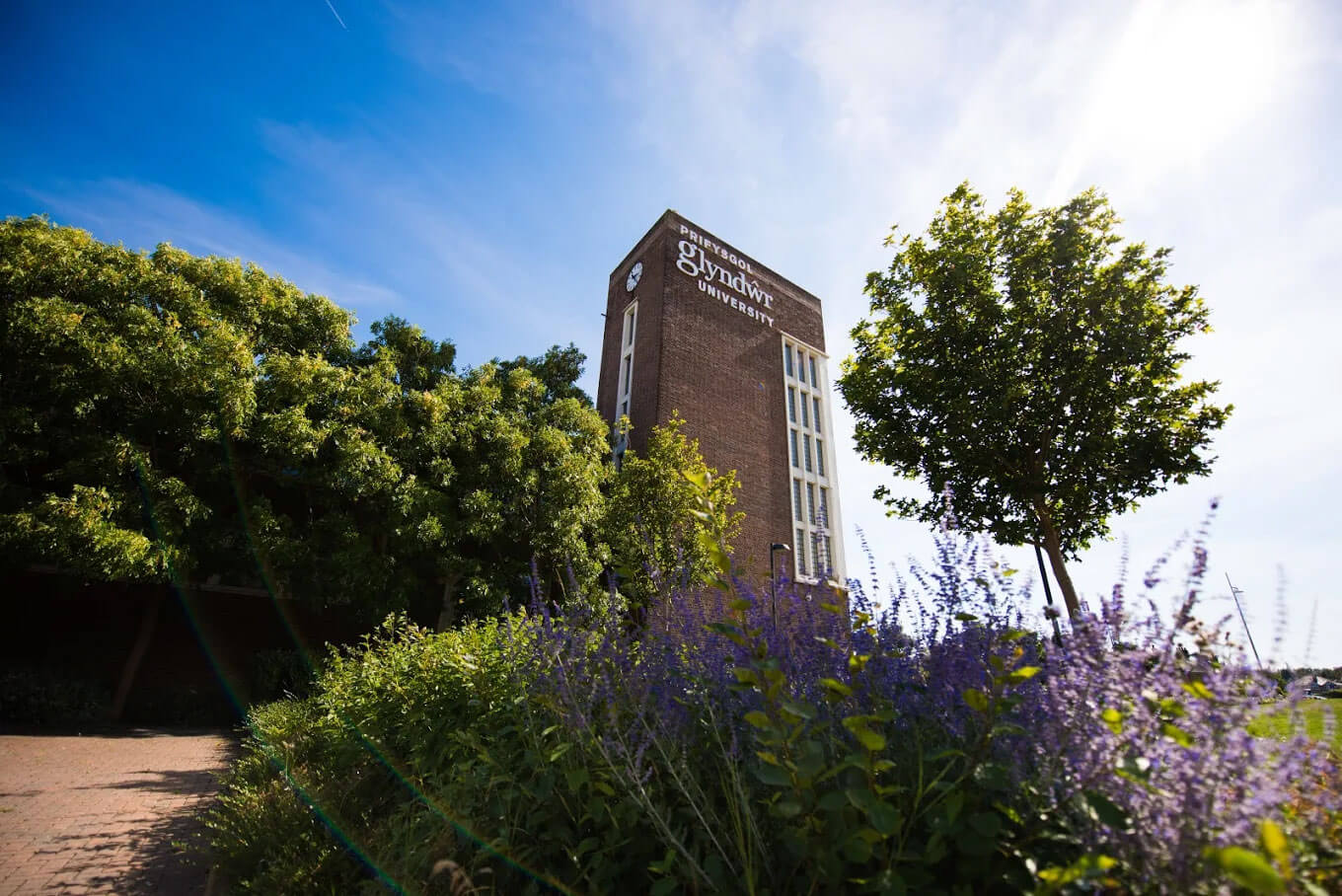 Image shows a Wrexham Glyndwr University building surrounded by trees and flowers and text that says Welcome to Living Life to the Full Wrexham Glyndwr University